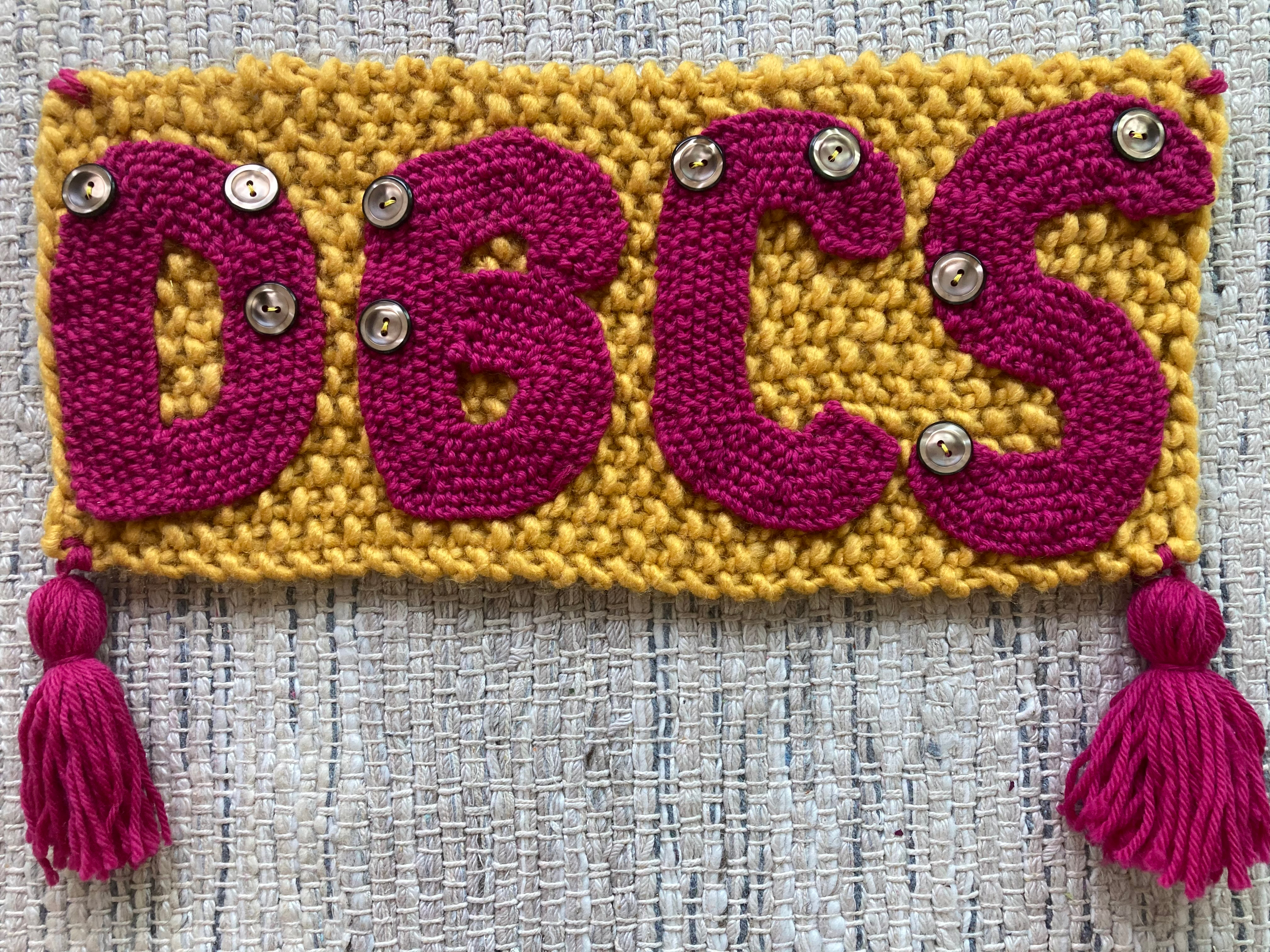 A knitted square of yarn with the text “DBCS” sewn on top. There are buttons sewn onto the letters to represent the braille for each letter.