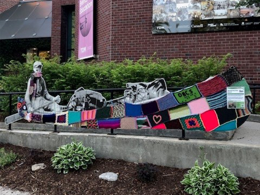 Tom Thompson’s canoe, covered In colourtul squares of knitted and crocheted yarn squares. One of the squares has the letters DBCS knitted onto it, with the braille underneath.