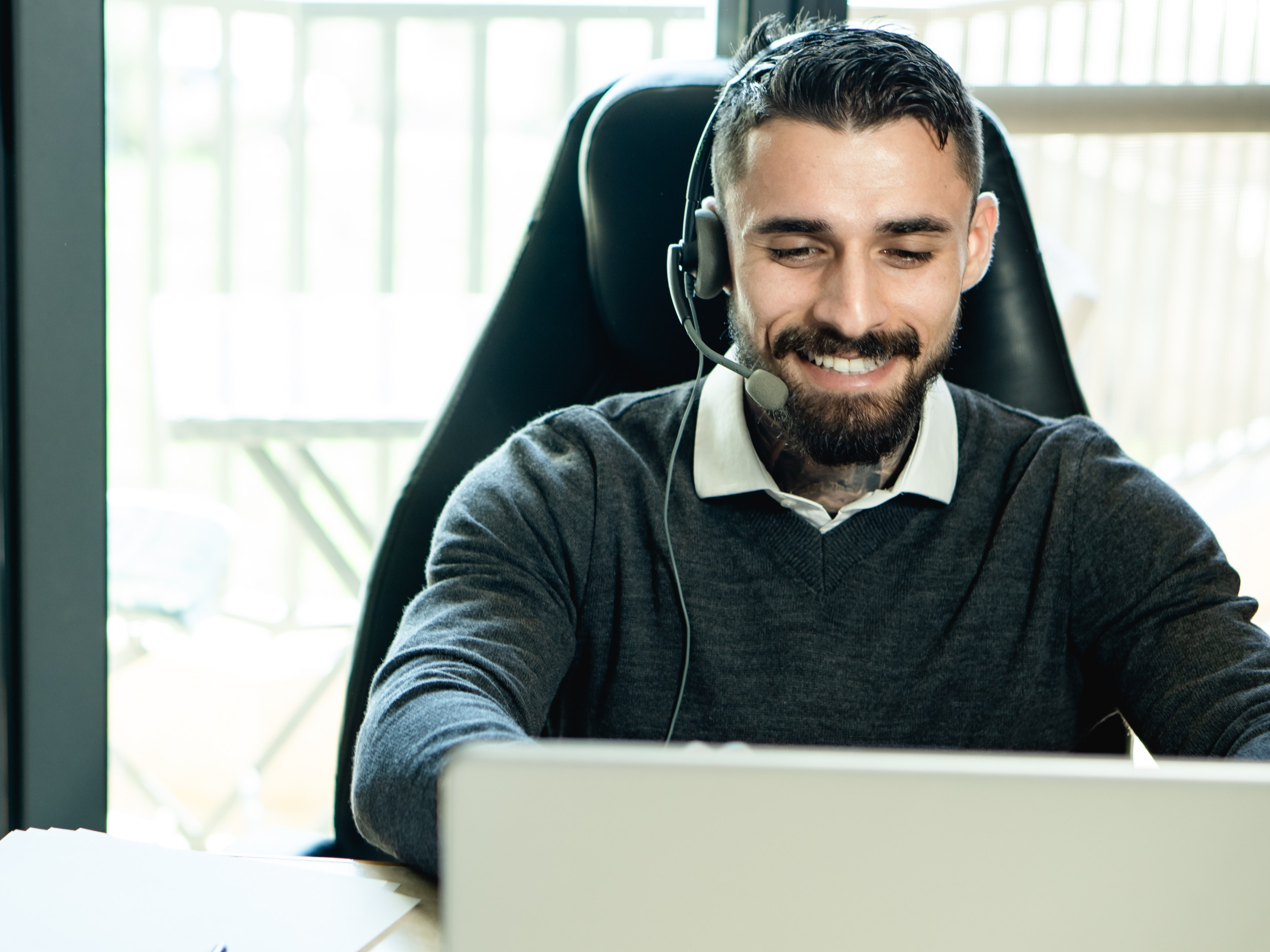 A man sits behind a computer screen, smiling while wearing a headset.