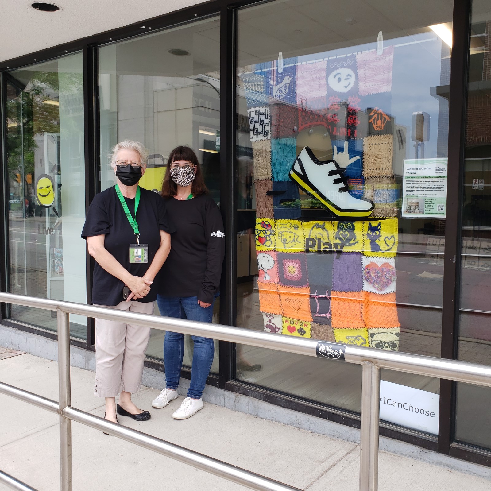 Two staff members stand outside a yarnbombing display in a window. 