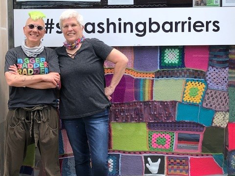 Two people pose In front of the CNIB GTA Hub building exterior. A collage/blanket of colourful yarn covers the main entryway window.