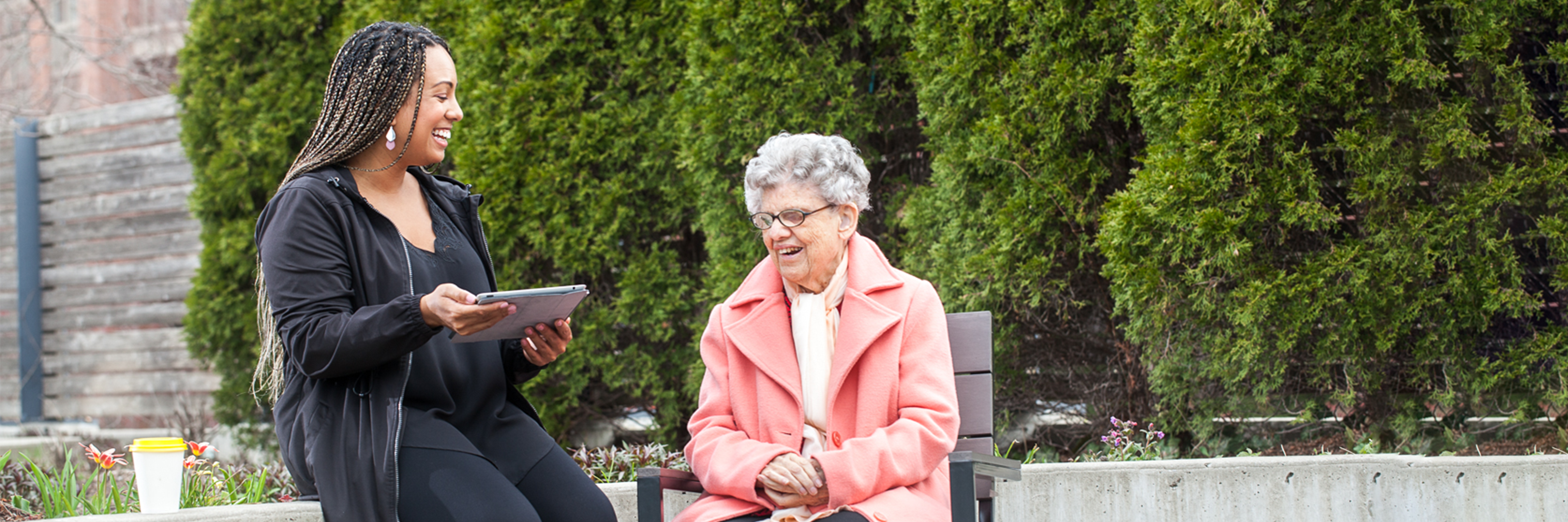 An intervenor and woman sit together outside with big smiles on their faces. The intervenor holds a tablet and types out a message using the client’s preferred communication method, large print notes.