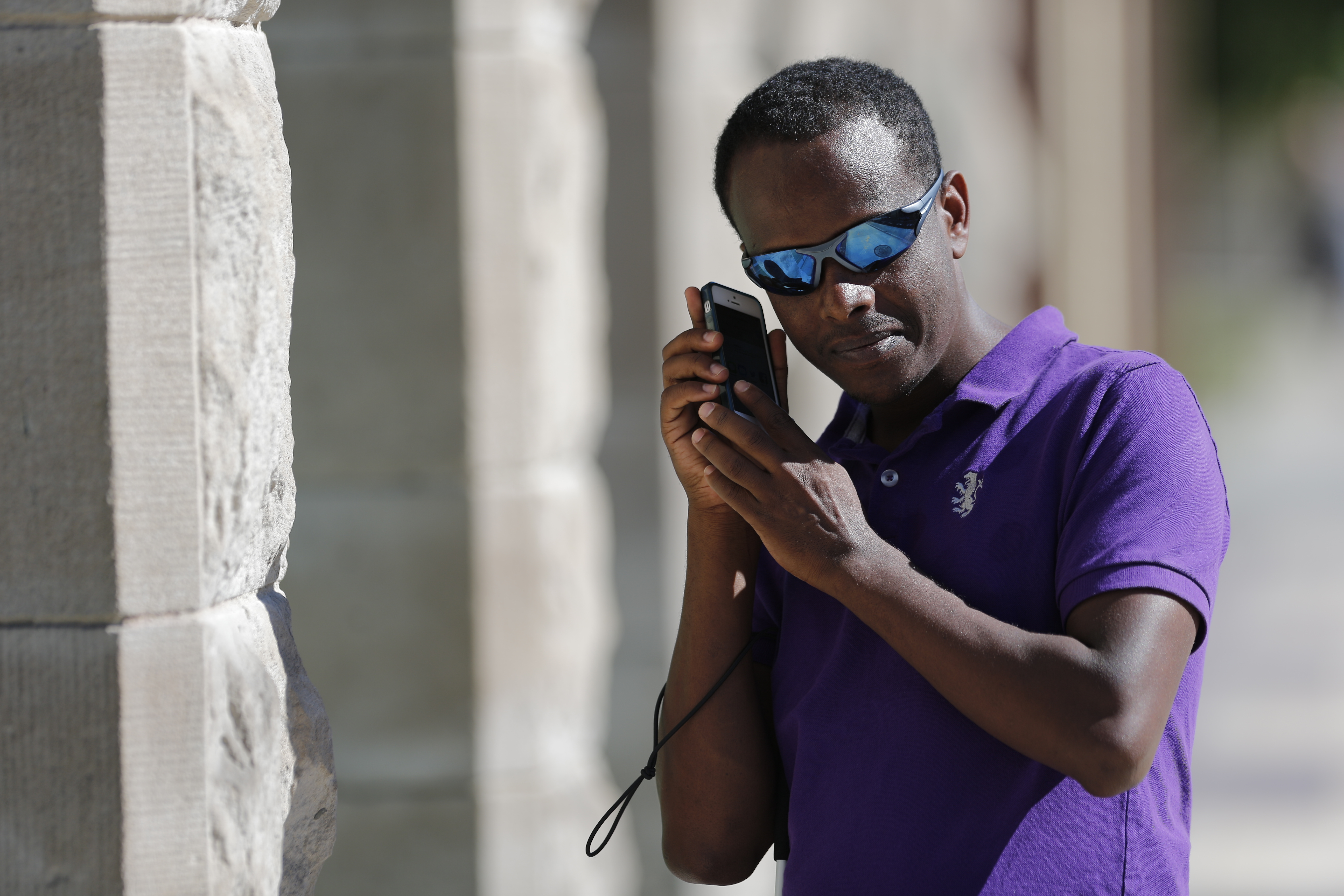 A man wearing sunglasses holds a smartphone up to his ear to listen. He holds a white cane under his arm. 