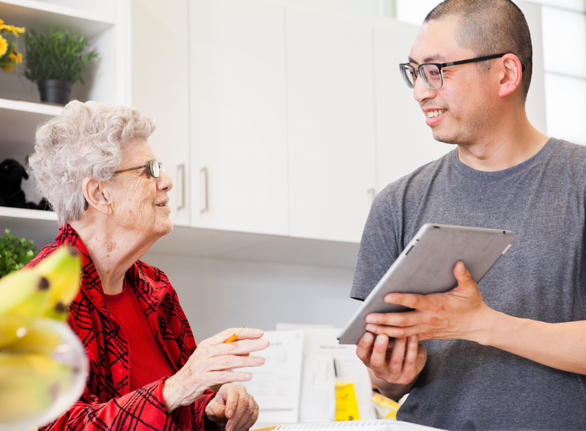 An intervenor holds up a tablet with large print notes for an older client to read. They are in a kitchen together. 