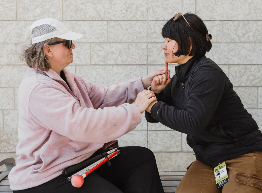 An intervenor uses tactile ASL to communicate with a person who is Deafblind. They are sitting together on a bench. 