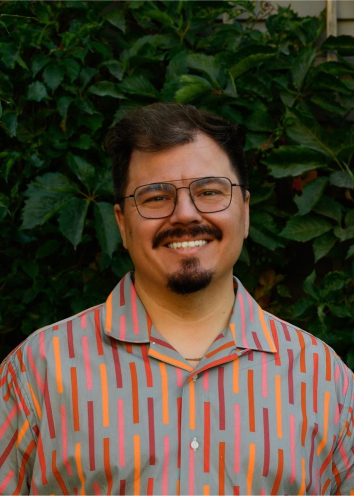 A smiling Dallas poses in front of greenery in an outdoor space. He wears a brightly coloured patterned shirt.