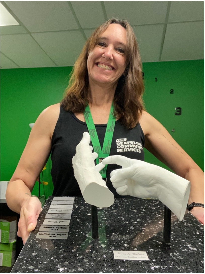Kelli-Ann smiles brightly while holding an award. She wears a CNIB Deafblind Community services top and lanyard.