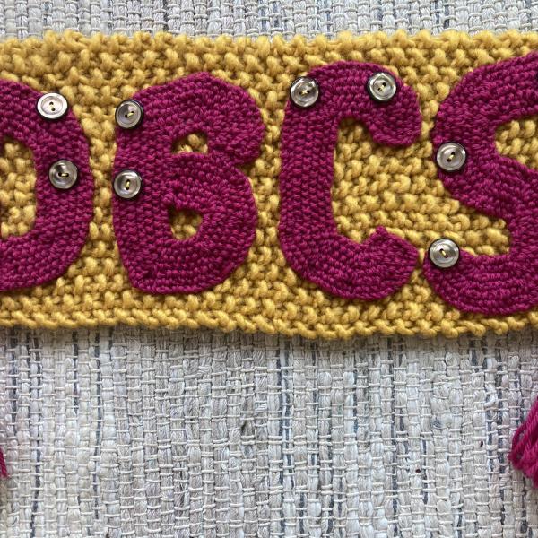 A knitted square of yarn with the text “DBCS” sewn on top. There are buttons sewn onto the letters to represent the braille for each letter.