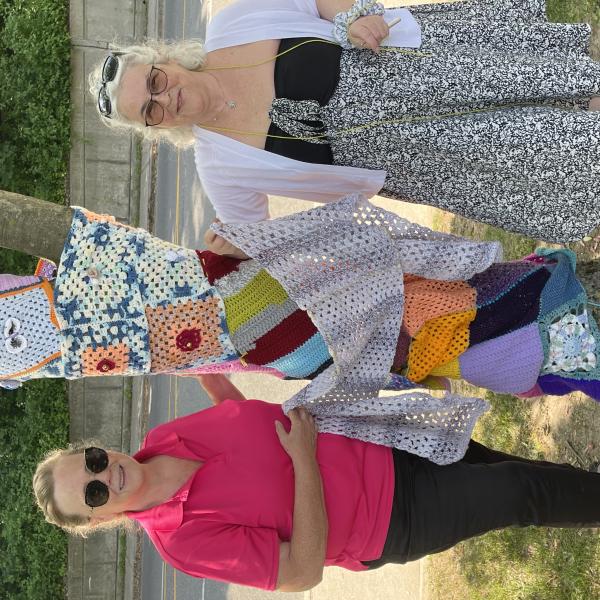 Two women stand in front of a tree covered in yarn. They are holding up an extra blanket of crocheted yarn.